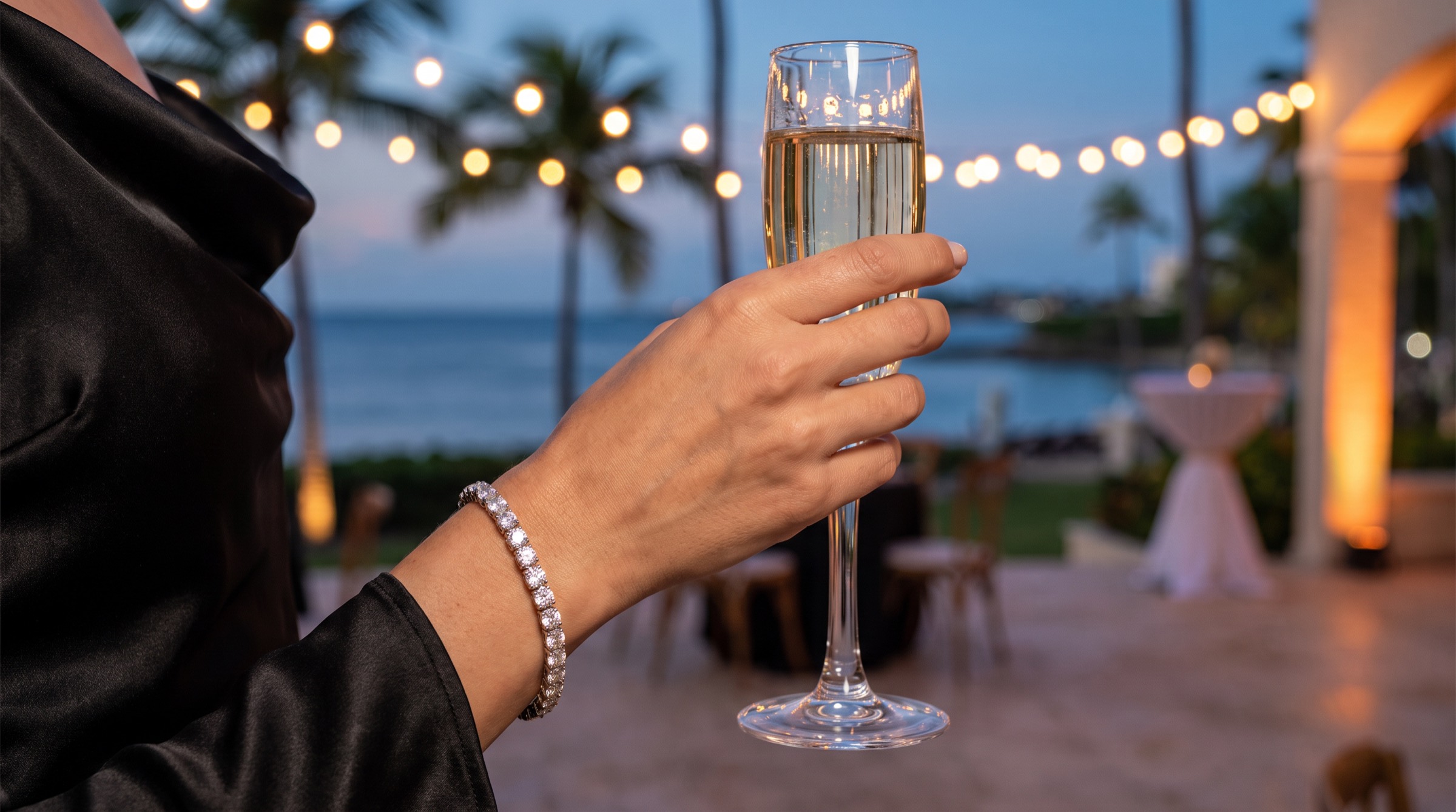 Mano de mujer con una pulsera tennis de diamantes sosteniendo una copa de champán en una terraza frente al mar